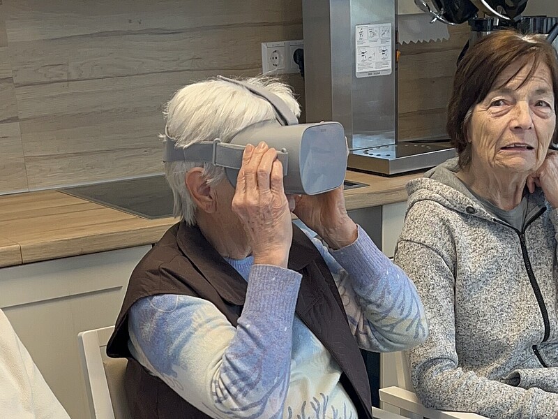 Eine alte Dame trägt eine VR-Brille, zwei alte Damen sitzen links und rechts daneben.
